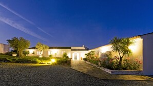 A well-lit residential building with a paved driveway leading to a gated entrance, surrounded by landscaped gardens and a clear night sky. - Monte Falperras (Mourão)