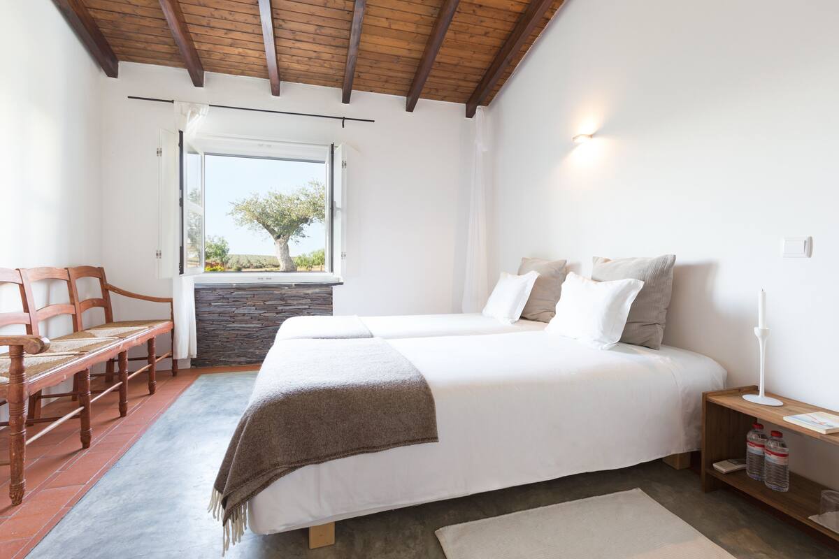 A bedroom with two beds, a wooden table, a bench, a stone wall, and a window with a view of trees.