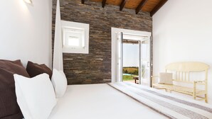 A bedroom with a stone wall, a bed with white bedding, a wooden bench, and a view of the outdoors. - Monte Falperras (Mourão)