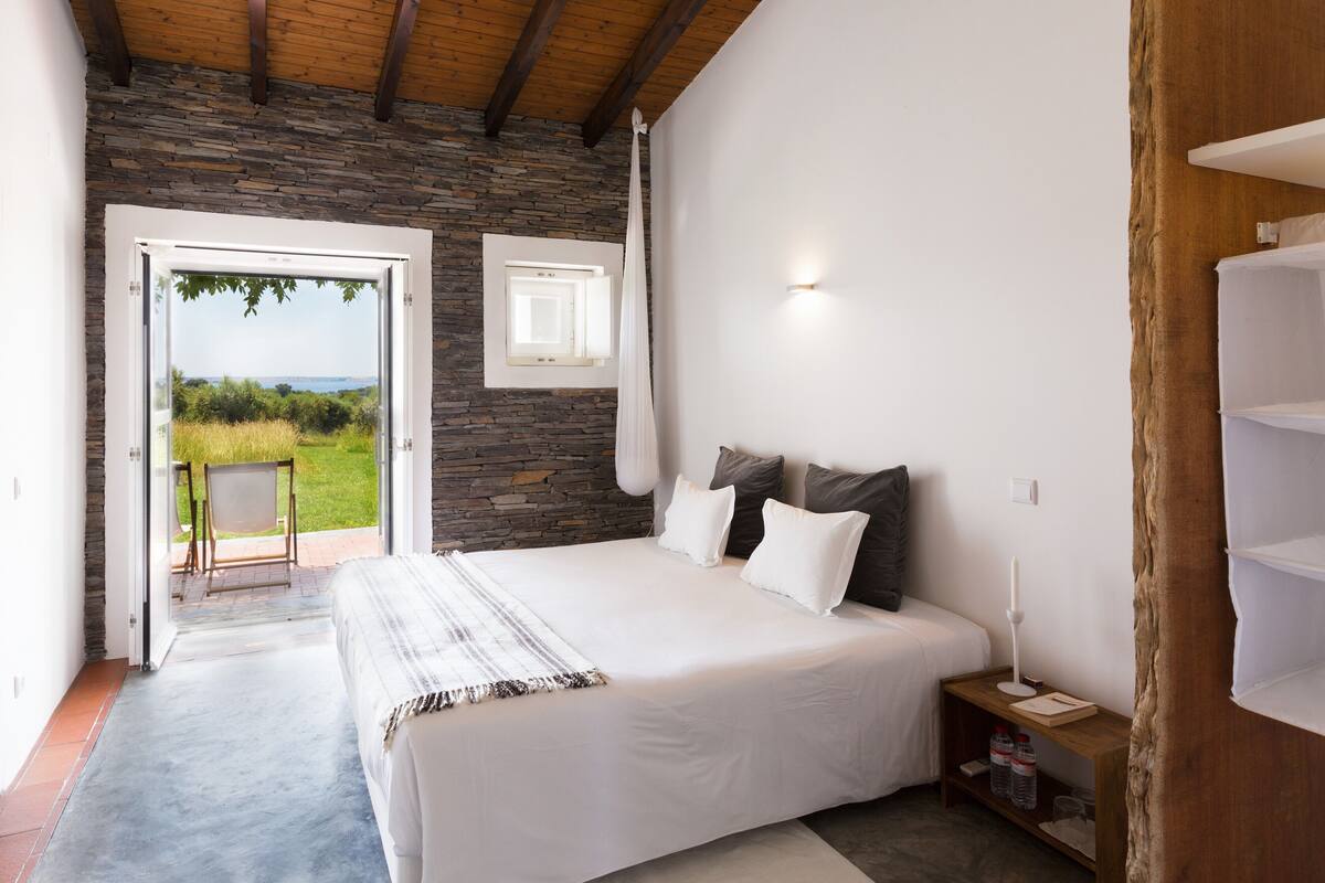 A bedroom with a large bed, a stone wall, a wooden ceiling, and a view of the outdoors.