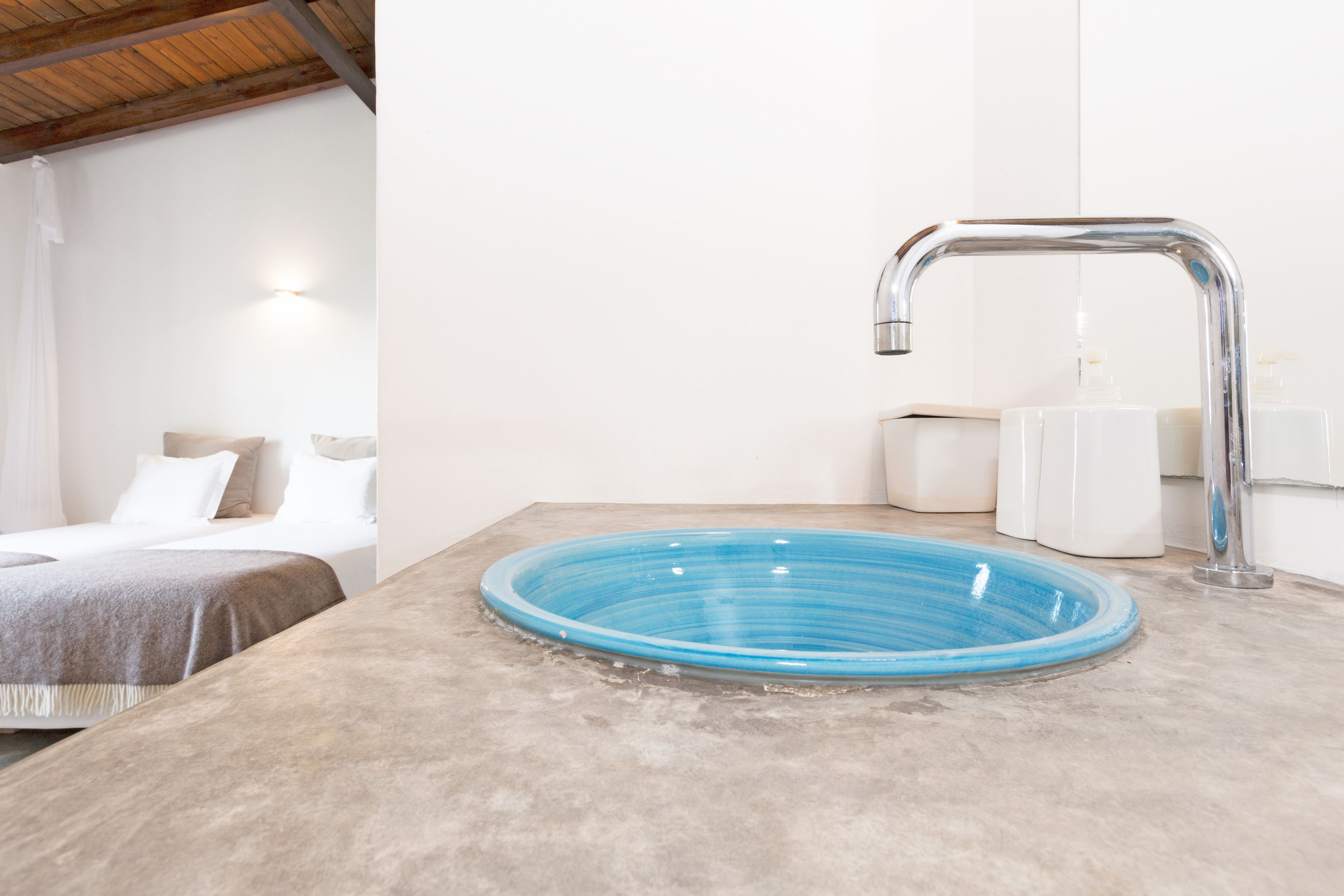 Un baño con un lavamanos de vidrio azul, un grifo moderno y una cama con ropa de cama blanca.