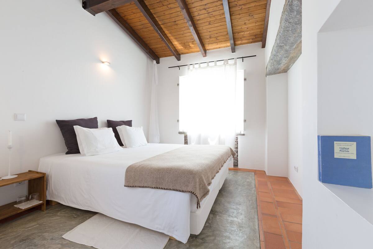 A bedroom with a large bed, a nightstand, a wall-mounted book, and a window with curtains.