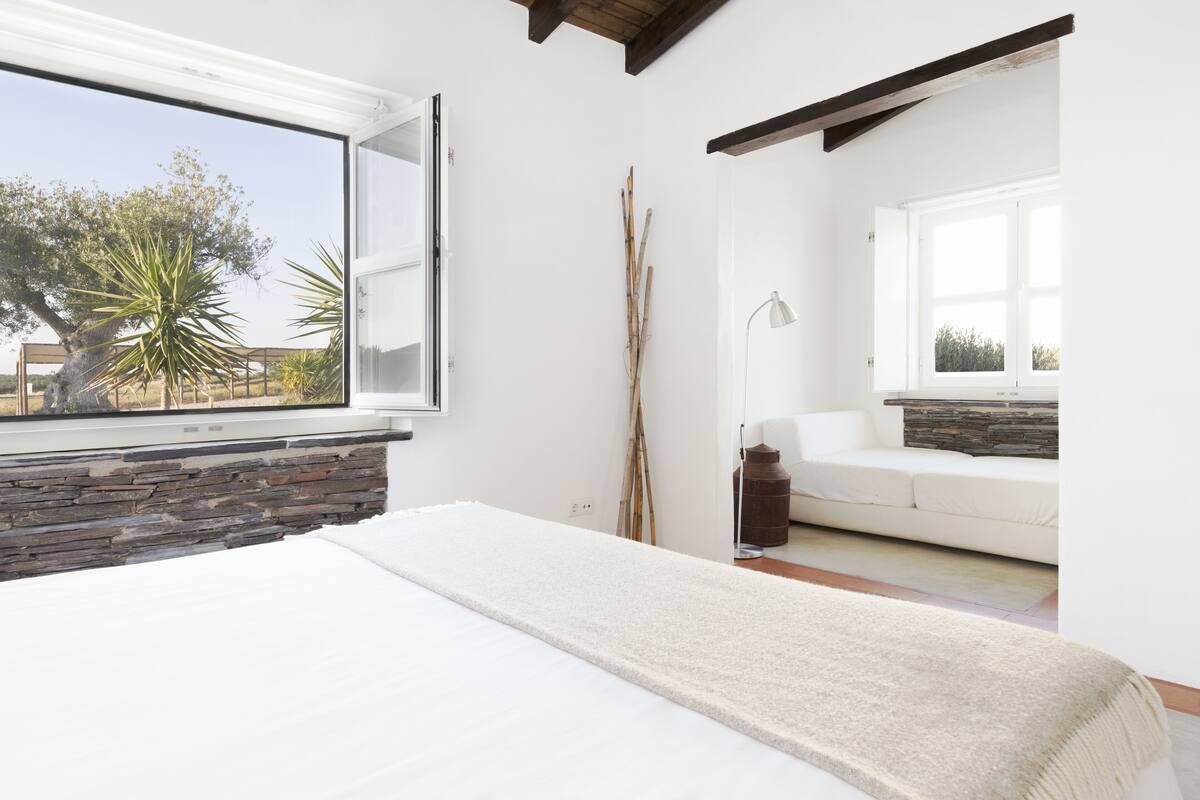 A bedroom with a large bed, a stone wall, a window with a view of trees, and a white sofa in the adjacent room.