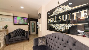 Un espace d’accueil d’hôtel avec un canapé de couleur sombre, un téléviseur fixé au mur et une enseigne indiquant « PERI SUITE ».