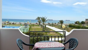 Een balkon met een tafel en stoelen, met uitzicht op een strand met palmbomen, een speeltuin en een helder uitzicht op de zee.