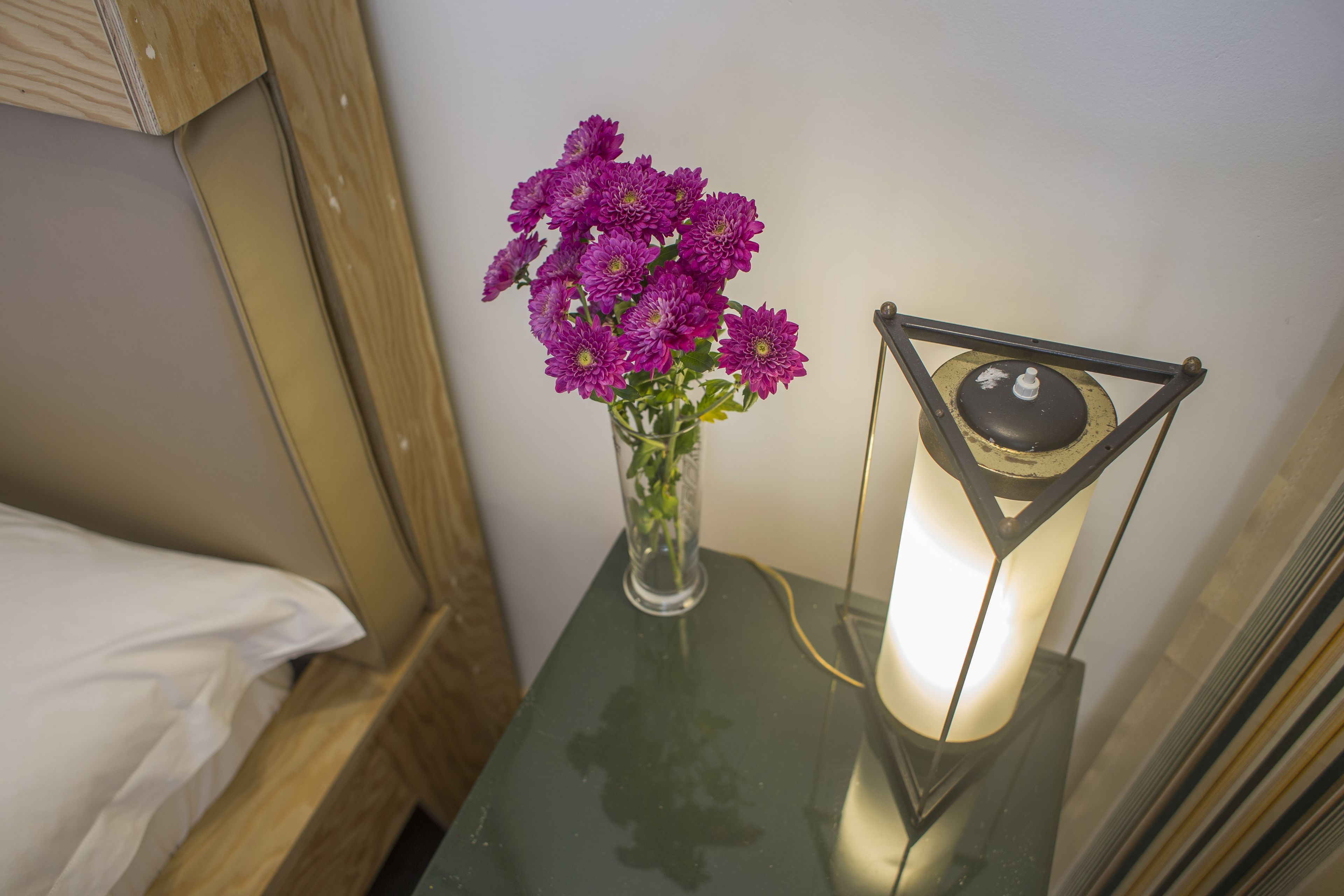 Ett litet bord med en vas med lila blommor och en vägglampa.