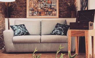 A grey sofa with two patterned pillows, a wooden side table, and a framed abstract artwork on the wall.
