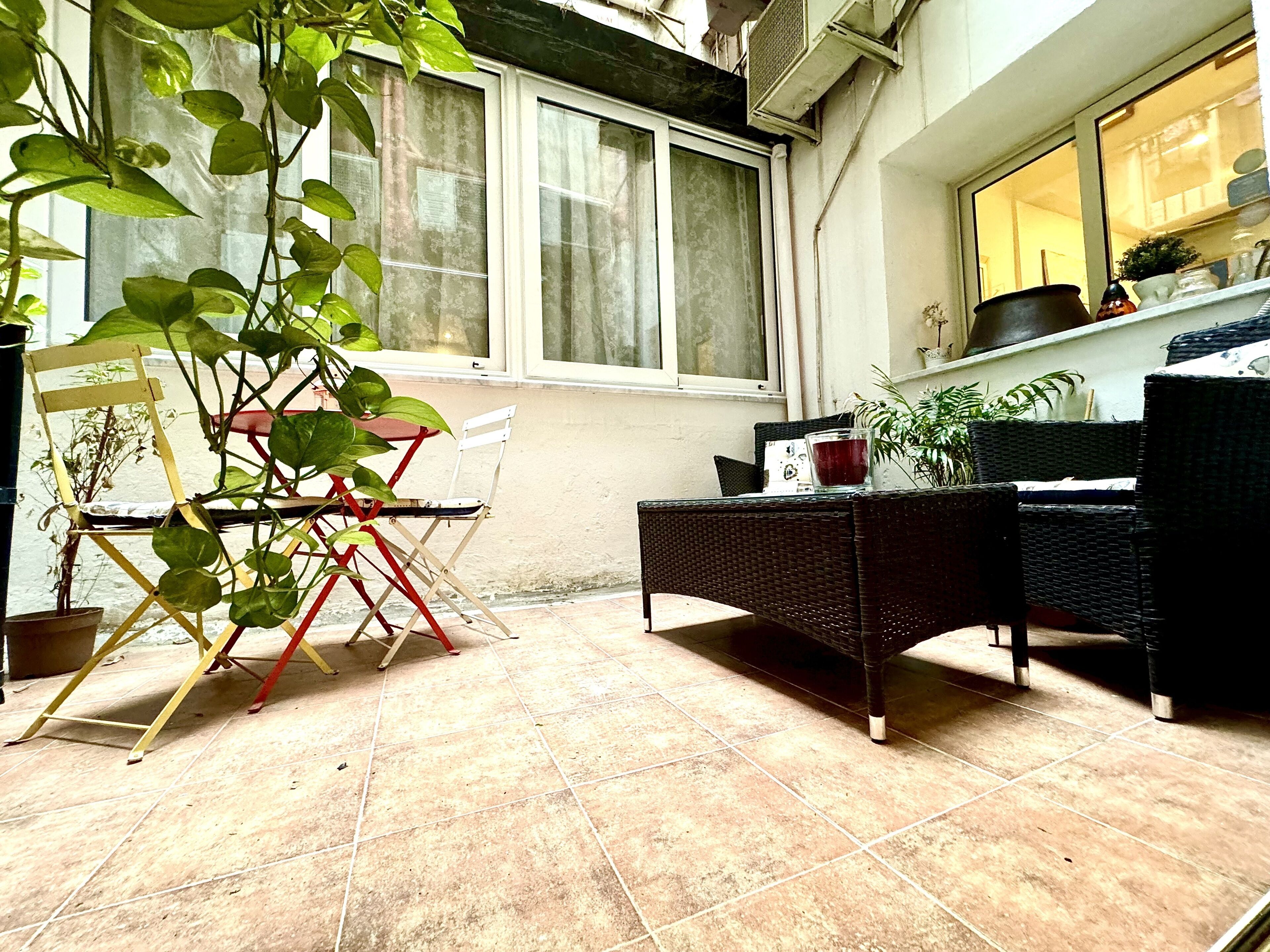 Un patio con una mesa y sillas de mimbre, una silla plegable y plantas en macetas.
