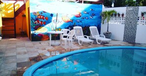 A swimming pool with a mural of fish and coral, white lounge chairs, and a tiled patio area. - Suítes São Francisco (Ipojuca)