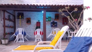 A patio with white plastic chairs and cushions, a tiled floor, and a building with a tiled roof and walls. - Suítes São Francisco (Ipojuca)