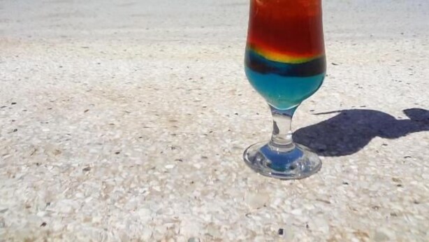A colorful drink with a tropical garnish on a beach with clear water and palm trees in the background. - Hôtel Keparanga (Mbour)