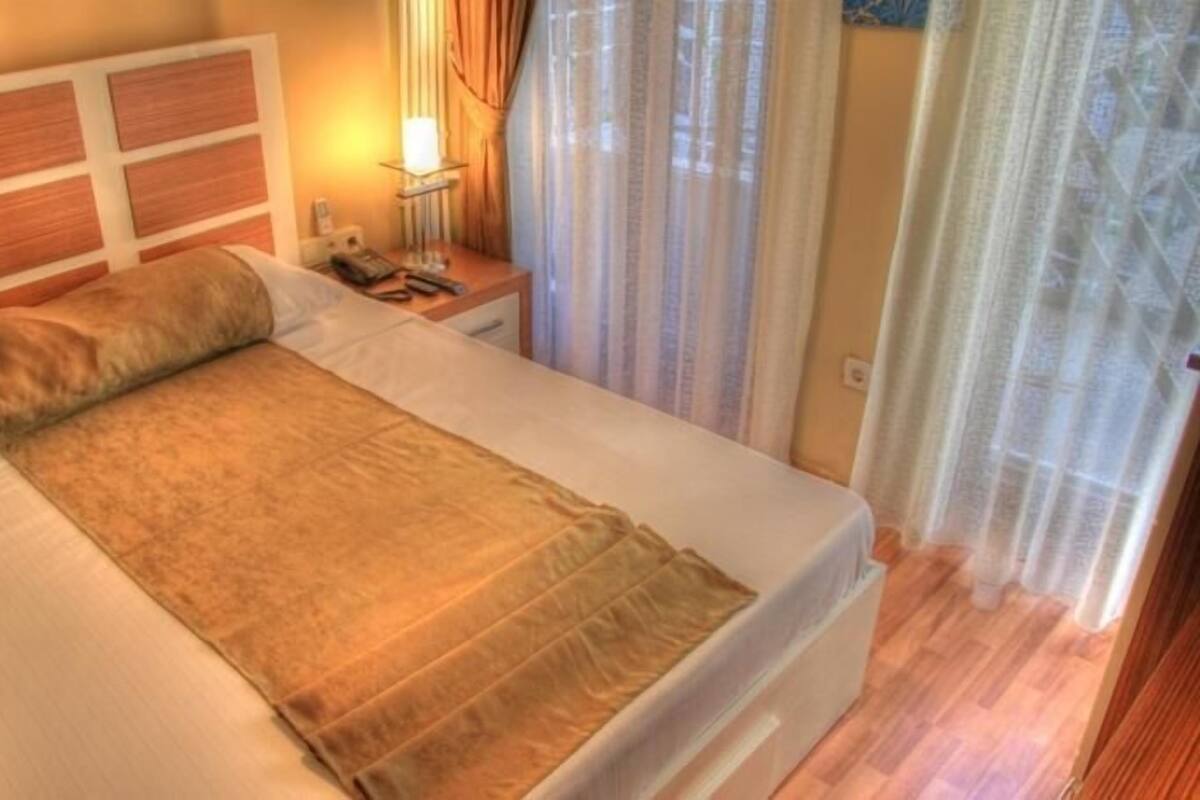 A neatly made bed with a headboard, a bedside table with a lamp, and a window with curtains.