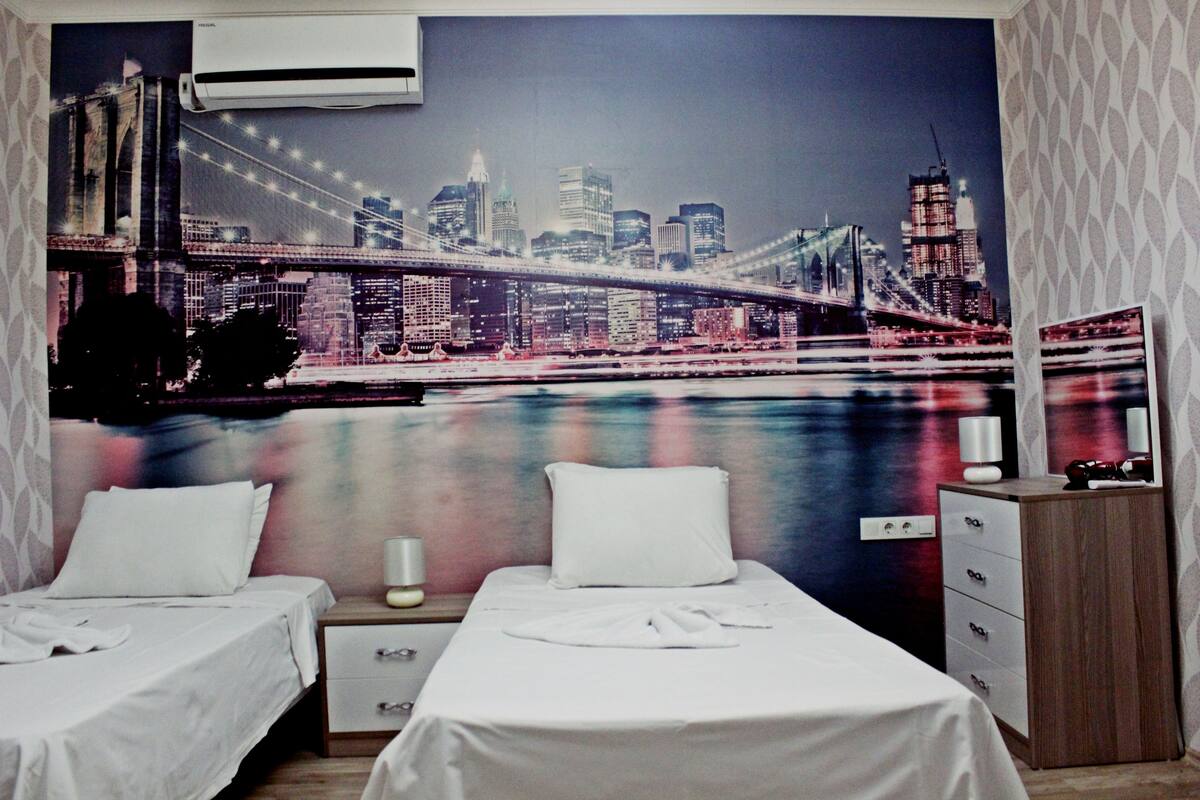 A hotel room with two beds, a nightstand, a dresser, and a large cityscape mural on the wall.