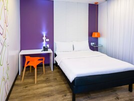 Quarto de hotel com cama, escrivaninha, cadeira, abajur e mesa de cabeceira.
