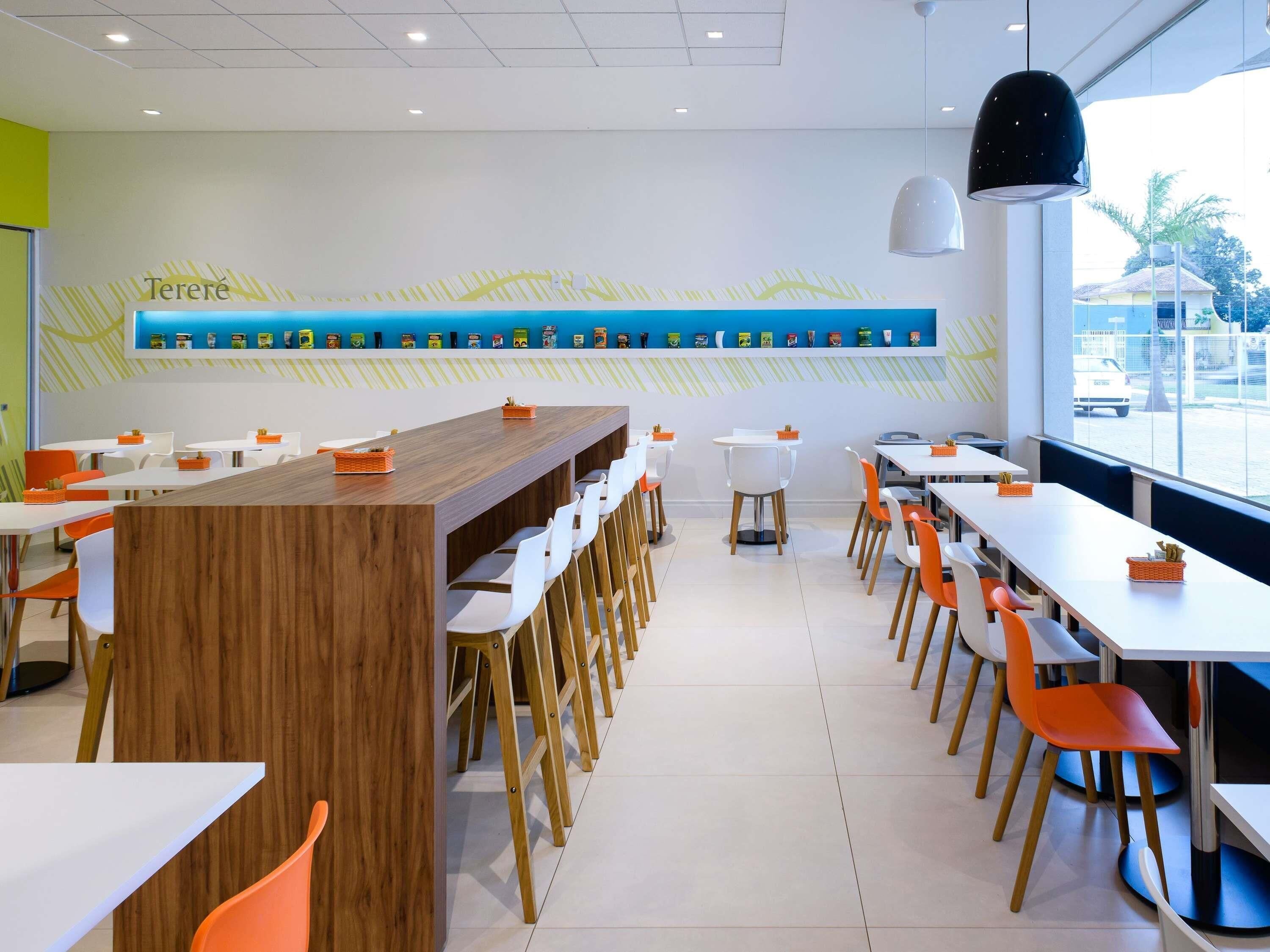 Interior moderno de restaurante con una larga barra de madera, sillas blancas y anaranjadas, y una exhibición de alimentos en la pared.
