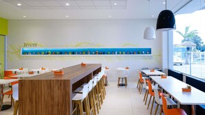 A modern restaurant interior with a long wooden counter, white and orange chairs, and a wall-mounted display of food items. - Ibis Styles Ponta Pora (Ponta Pora)