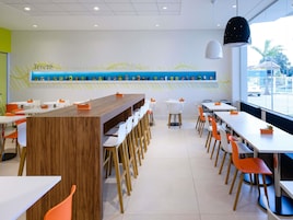 Um restaurante moderno com um balcão de madeira comprido, cadeiras brancas e laranja, e uma vitrine de comida na parede.