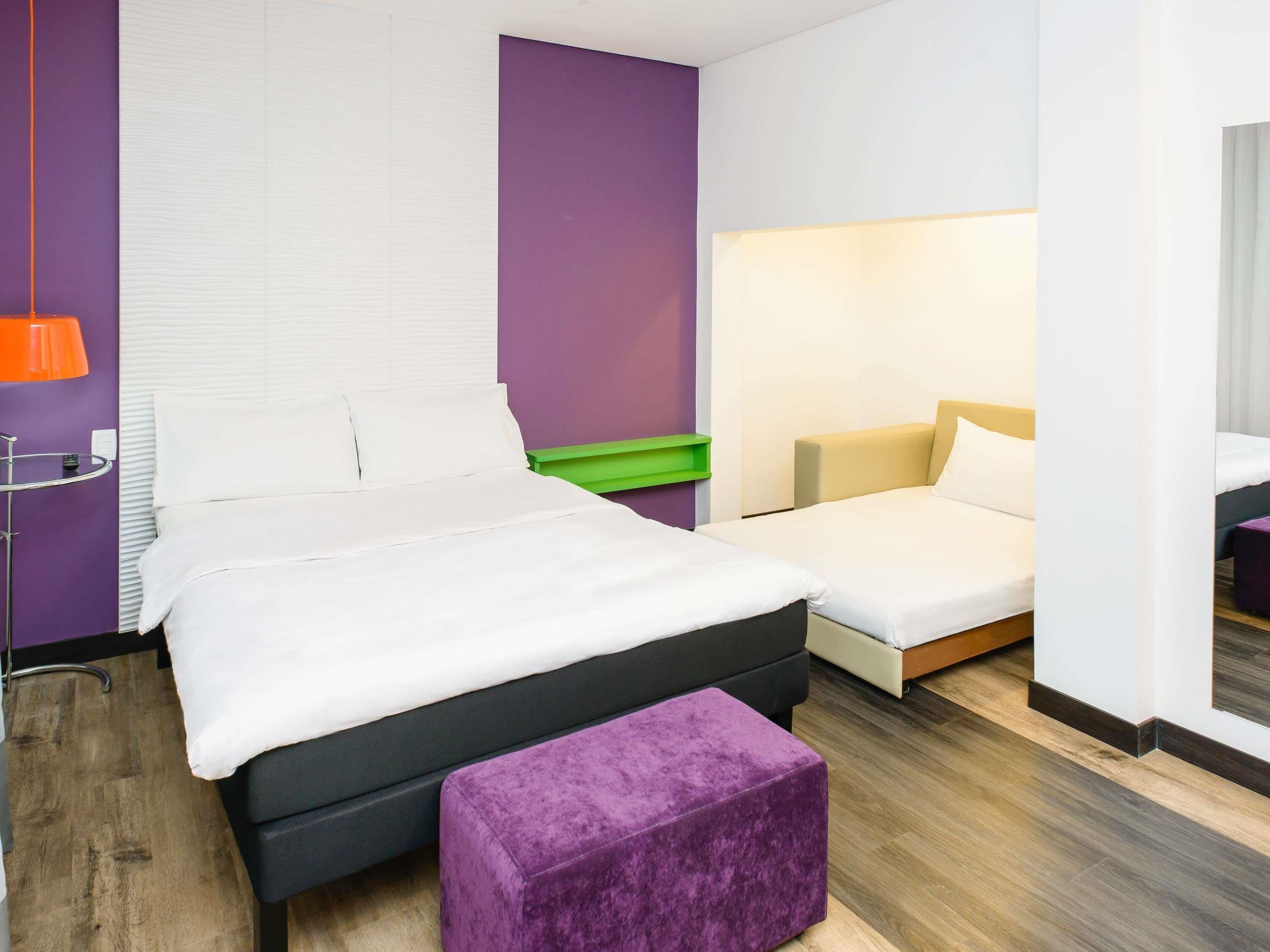 A hotel room with a large bed, a smaller bed, a purple ottoman, a green nightstand, and a wall-mounted lamp.
