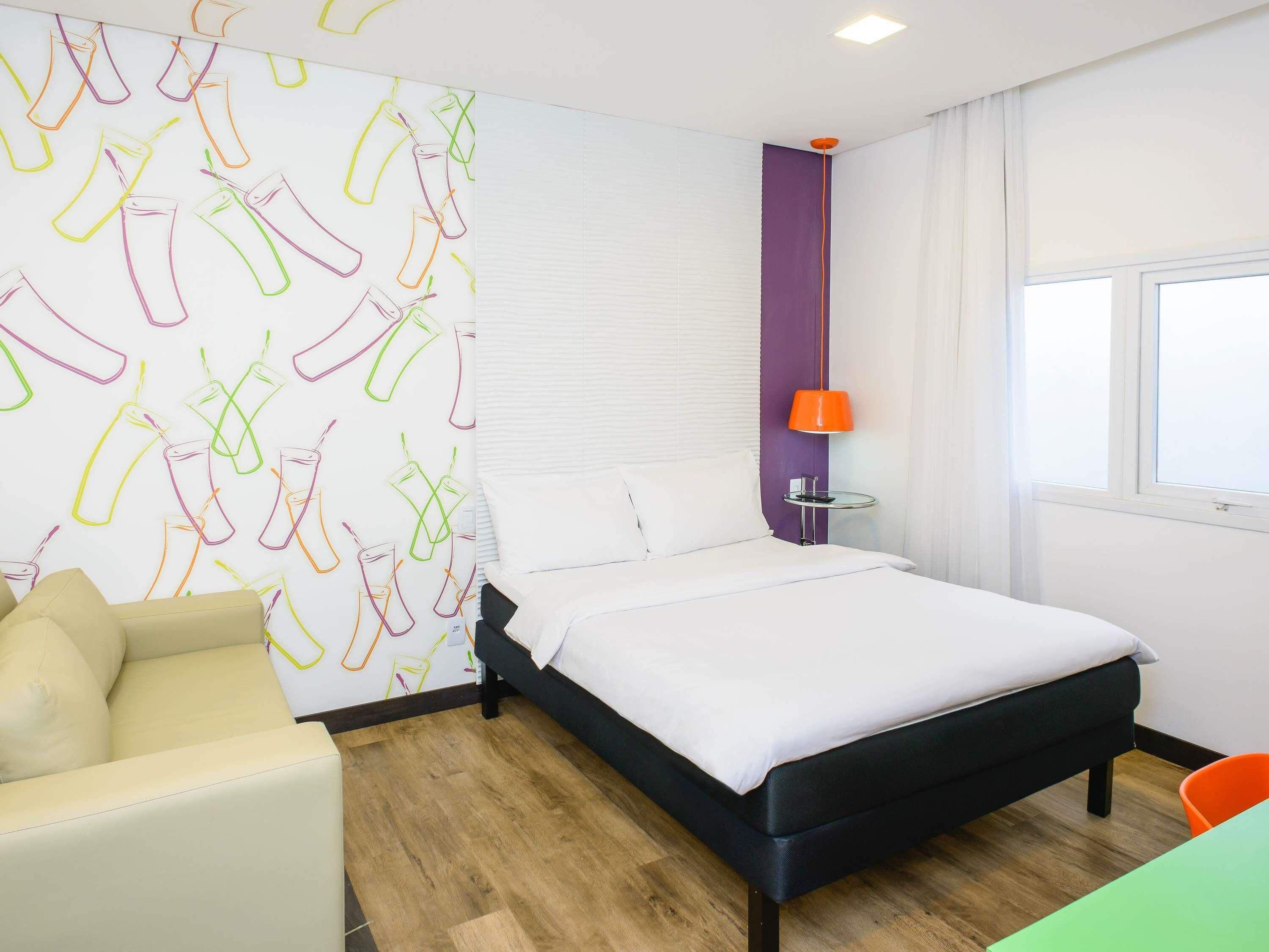 A hotel room with a bed, a chair, a table, a lamp, and a window.