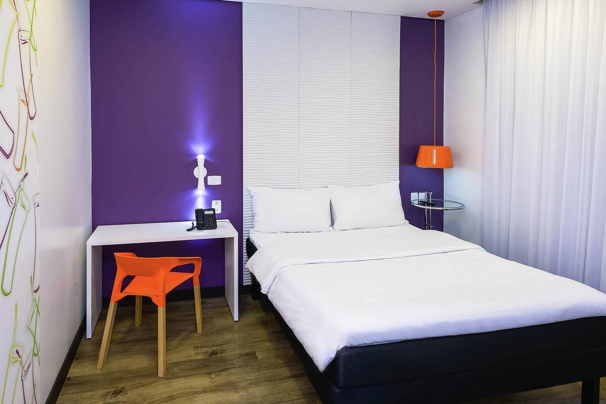 A hotel room with a bed, a desk, a chair, a phone, a lamp, and a window with blinds.