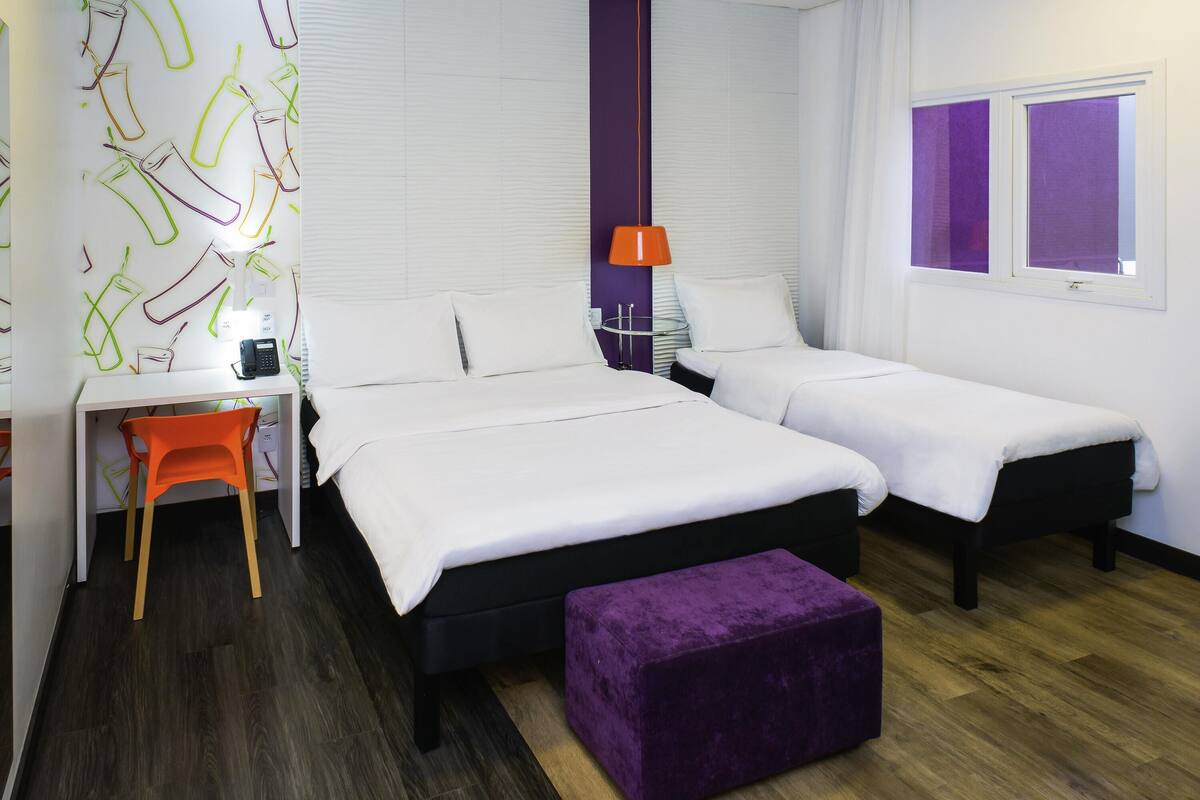 A hotel room with two beds, a desk with a chair, a purple ottoman, and a window with blinds.