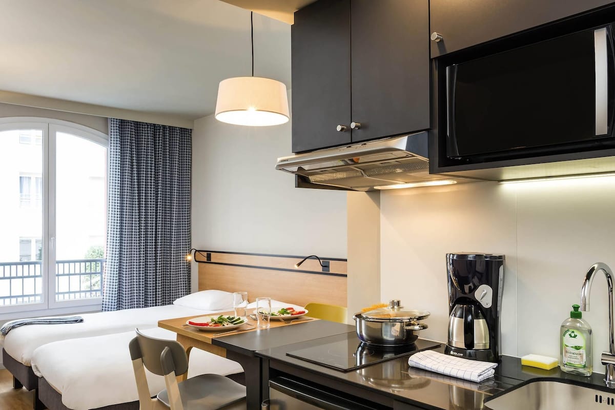 Ein modernes Hotelzimmer mit Küchenzeile, einem Bett, einem Esstisch mit Frühstück und einem großen Fenster.