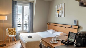A hotel room with a bed, a desk with a coffee maker, a chair, a lamp, and a view of buildings through the window. - Aparthotel Adagio Access La Défense Puteaux (Puteaux)