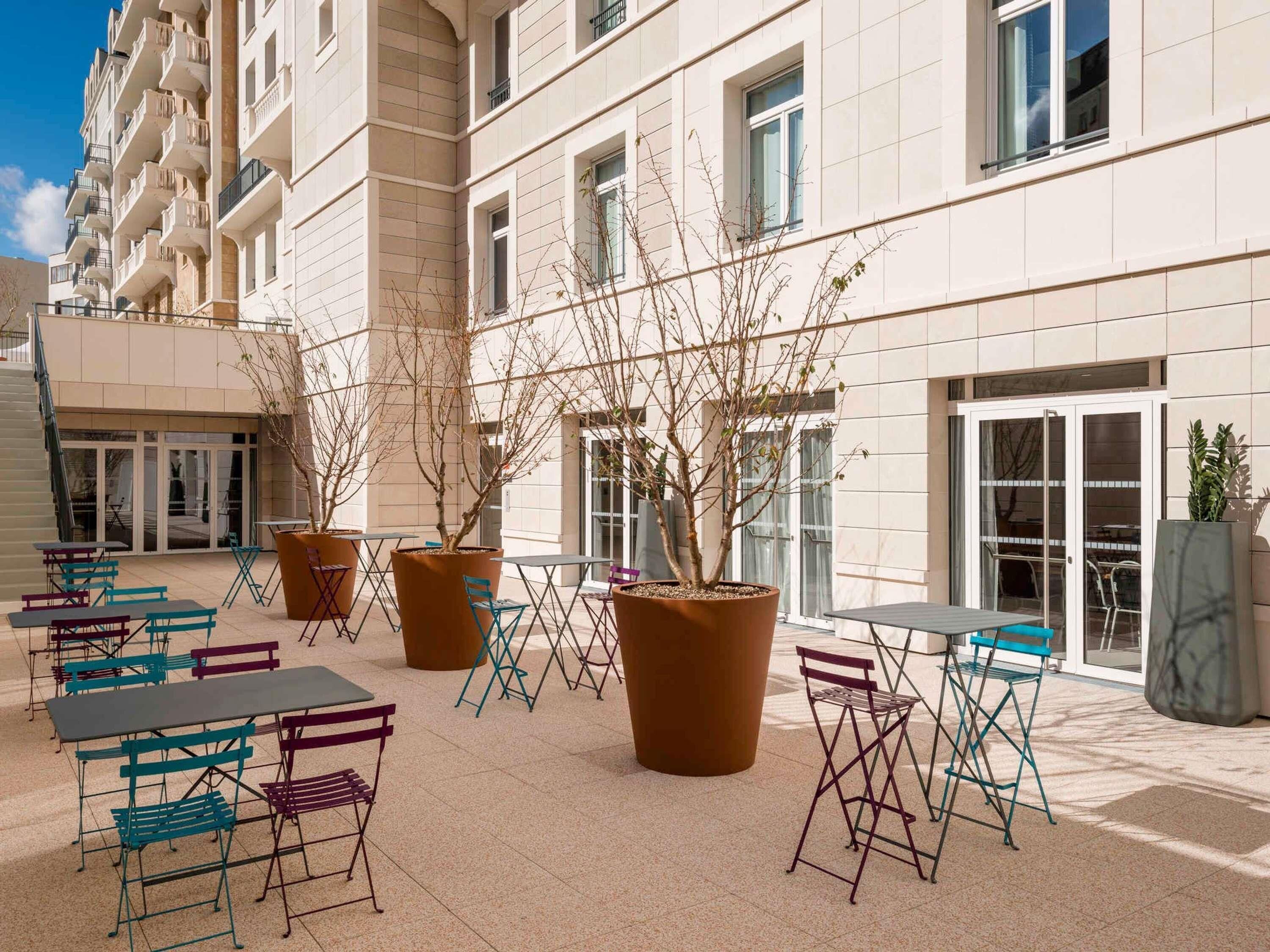 An outdoor patio area with multiple tables and chairs, potted plants, and a building facade with balconies and windows.