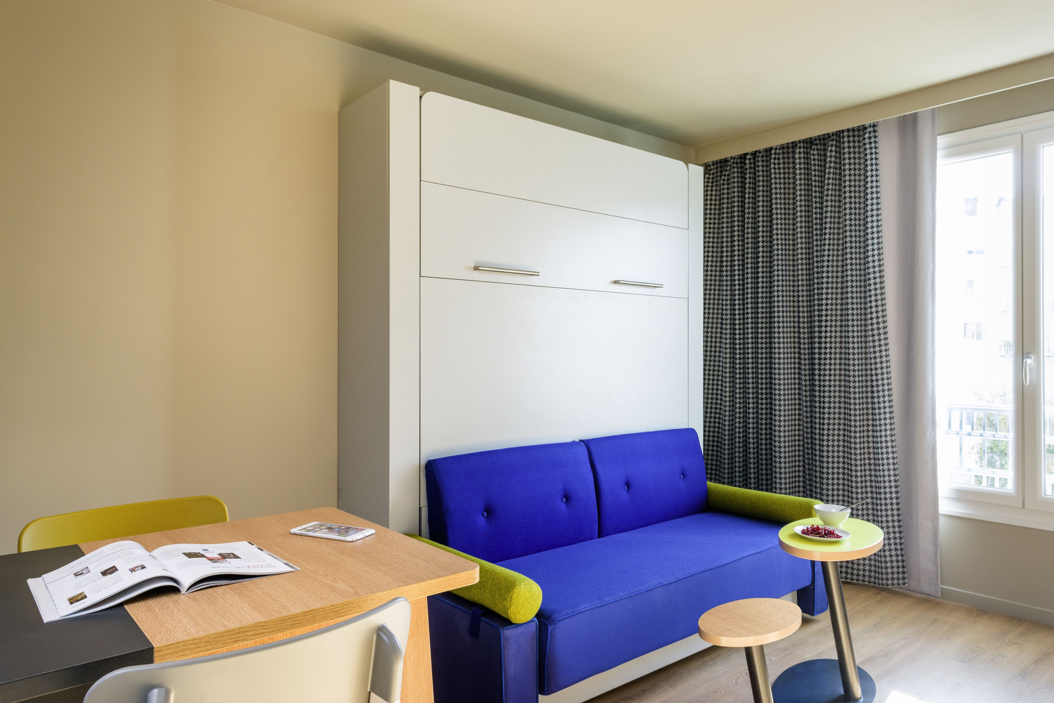 A room with a blue sofa, a wooden table with a magazine, a small round table with a bowl, and a window with curtains.