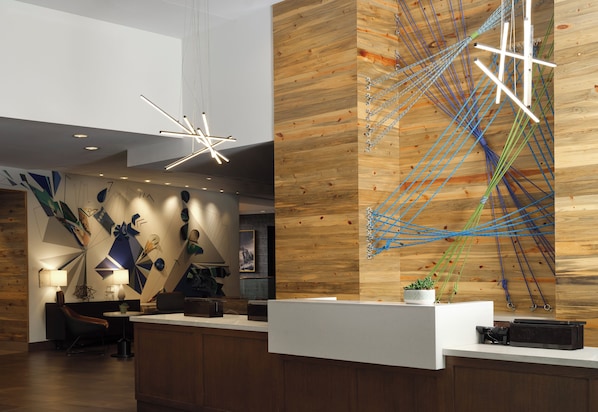 A modern reception area with a wooden wall, abstract art, a white reception desk, and a unique ceiling light fixture. - Hyatt Place Denver Downtown (Denver)