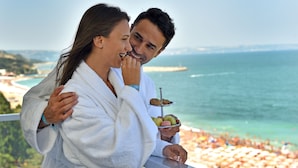 A couple in white robes overlooking a beach with a clear view of the ocean and a distant pier.