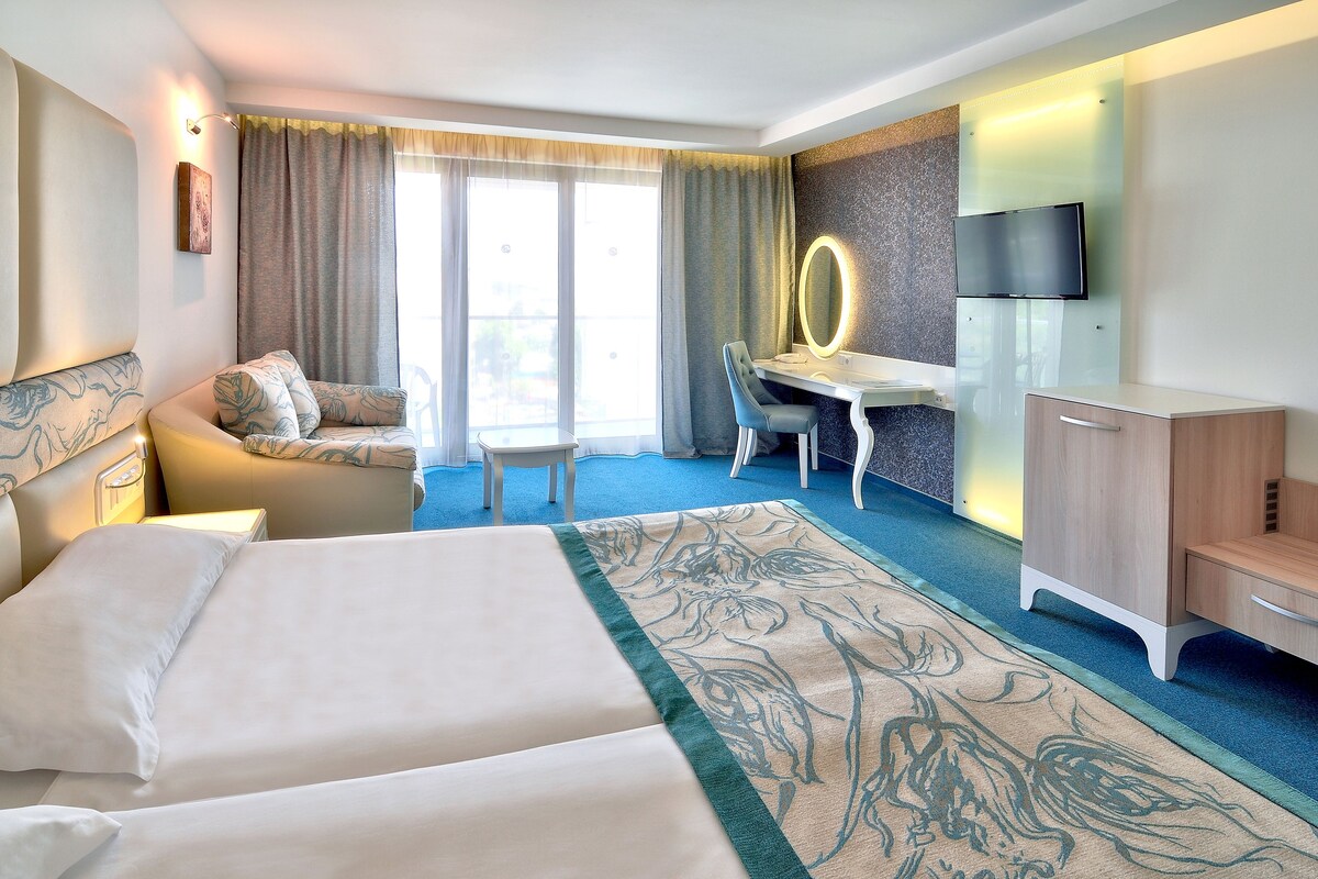 A hotel room with a large bed, a desk with a mirror, a TV, and a seating area with a sofa and chairs.
