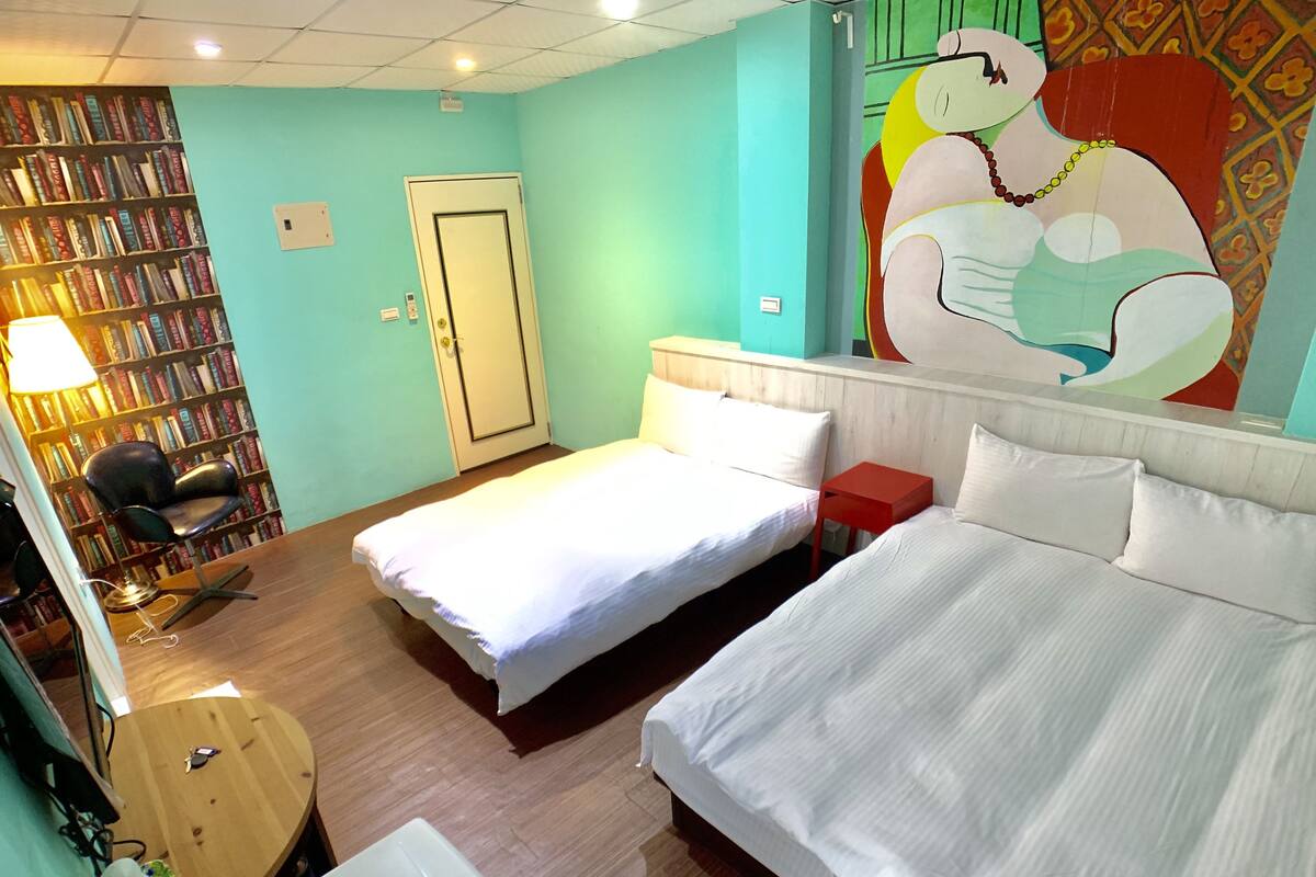 A hotel room with two beds, a desk, a chair, a bookshelf, and a door.