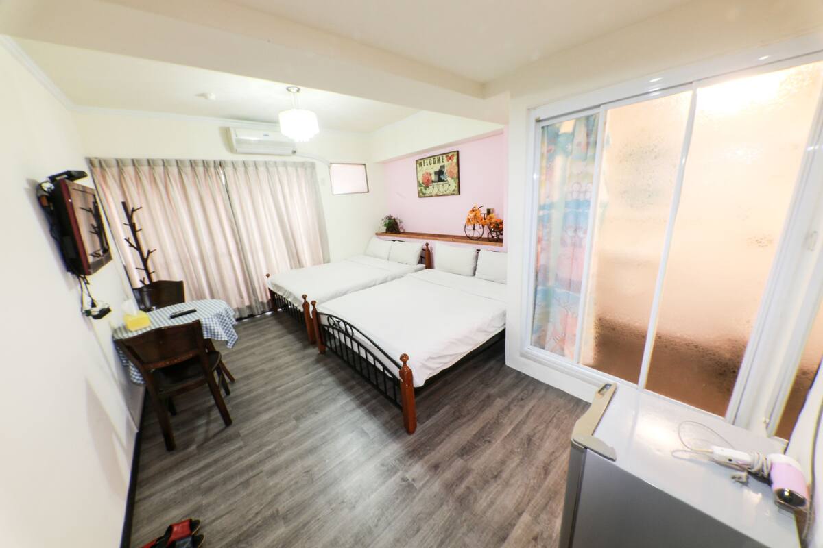 A bedroom with two beds, a desk, a chair, a window with frosted glass, and a wall-mounted air conditioner.