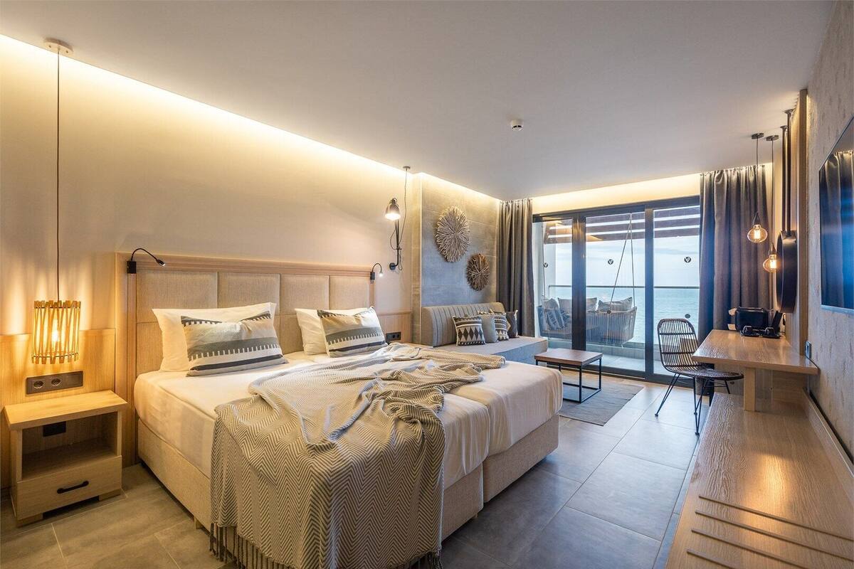 A modern bedroom with a large bed, a desk, and a balcony with a view.