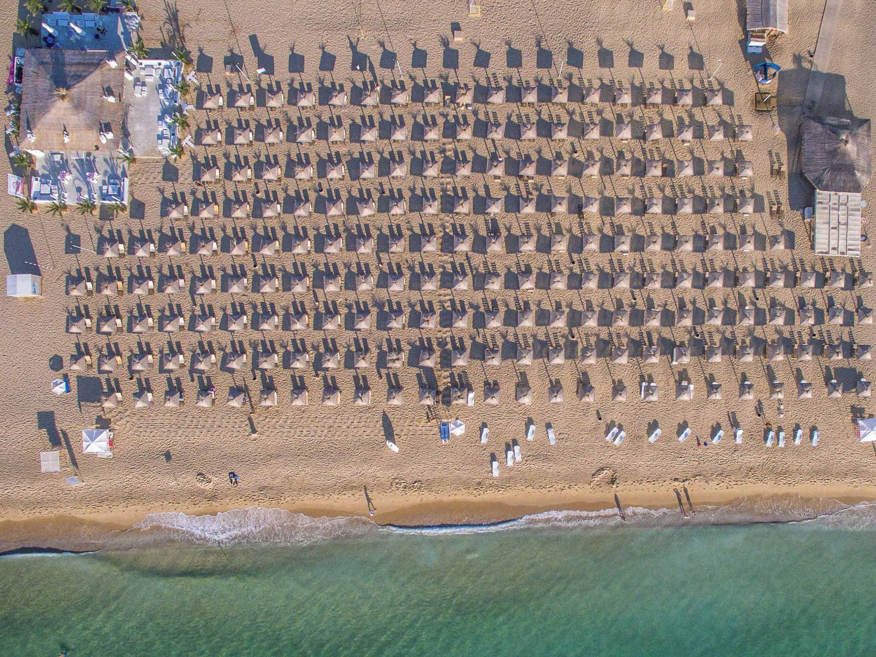 An aerial view of a beach with rows of umbrellas and beach chairs, a few people, and a building with a pool in the background.