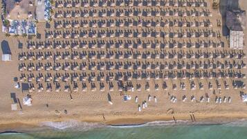 An aerial view of a beach with rows of umbrellas and beach chairs, a few people, and a building with a pool in the background.