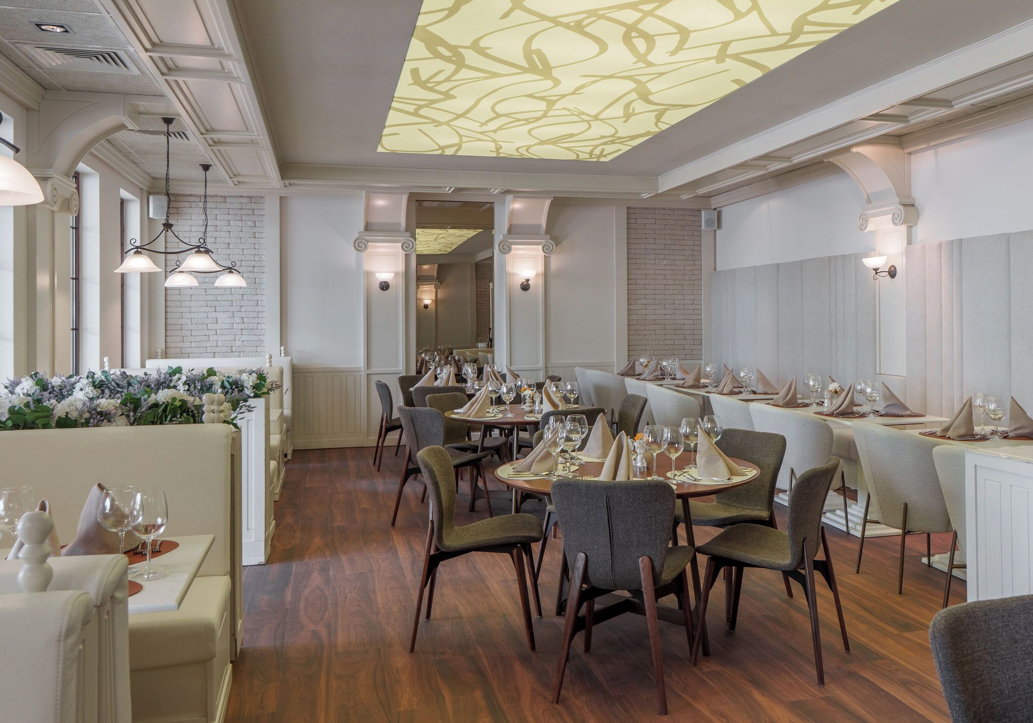 A restaurant with wooden flooring, white walls, and a ceiling with a light pattern. Tables are set with wine glasses and plates.