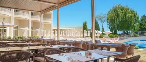 A covered outdoor dining area with tables and chairs, a pool, and a view of the surrounding landscape.