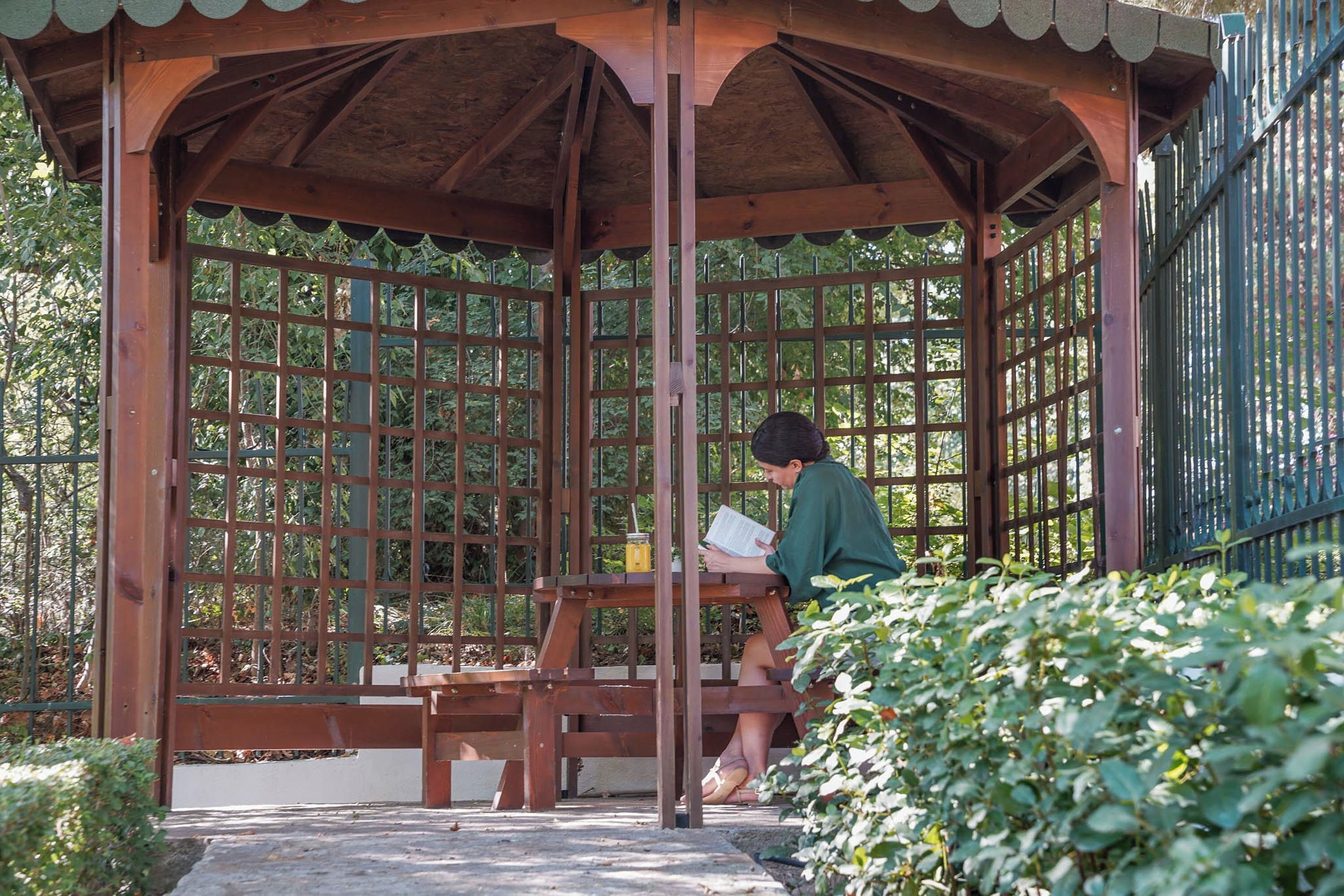 A wooden gazebo with a lattice design, a bench, and a person reading a book.