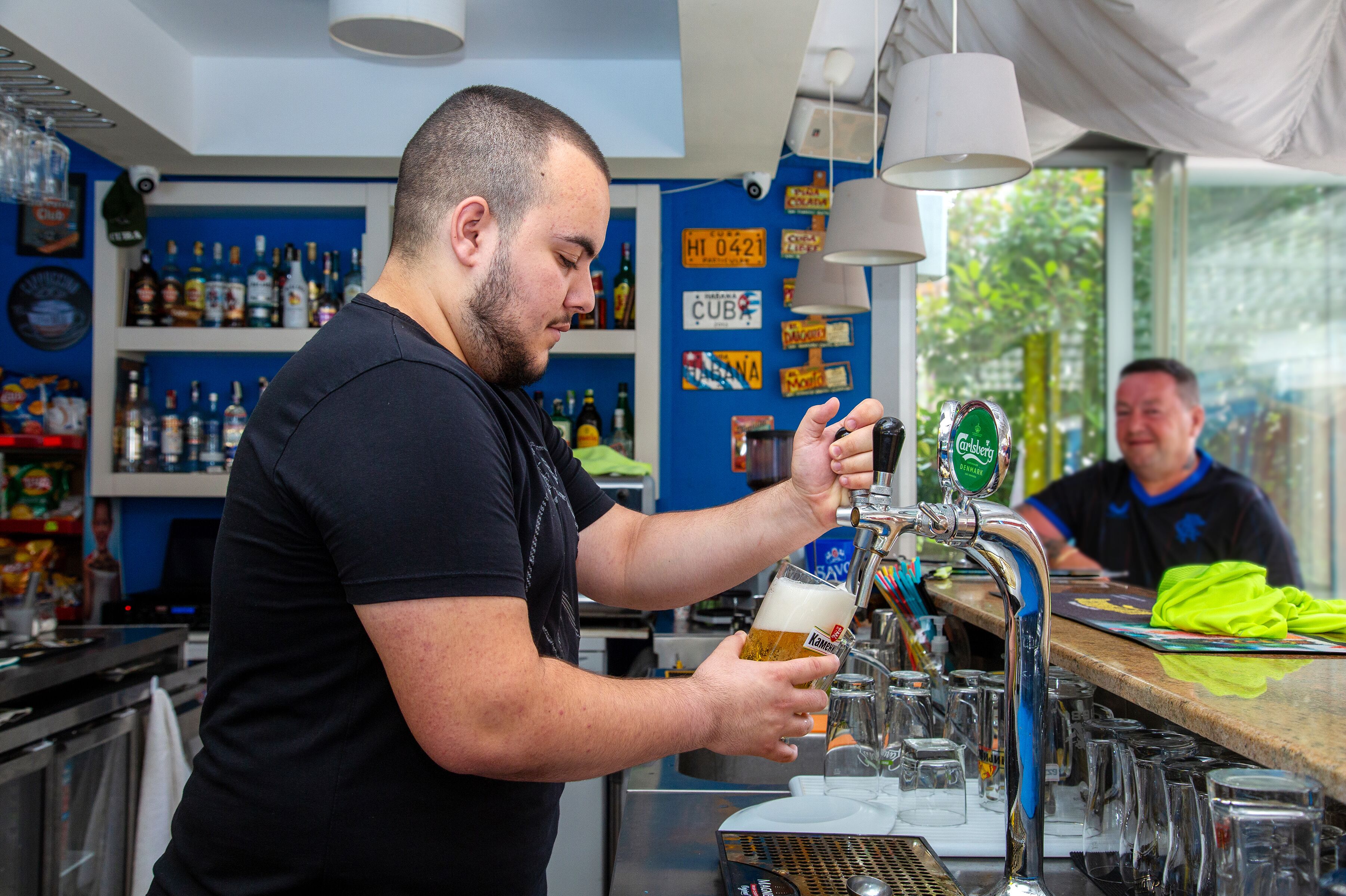 Un barman verse de la bière dans un verre au comptoir d’un bar, où l’on peut voir des étagères remplies de bouteilles et divers panneaux accrochés au mur.