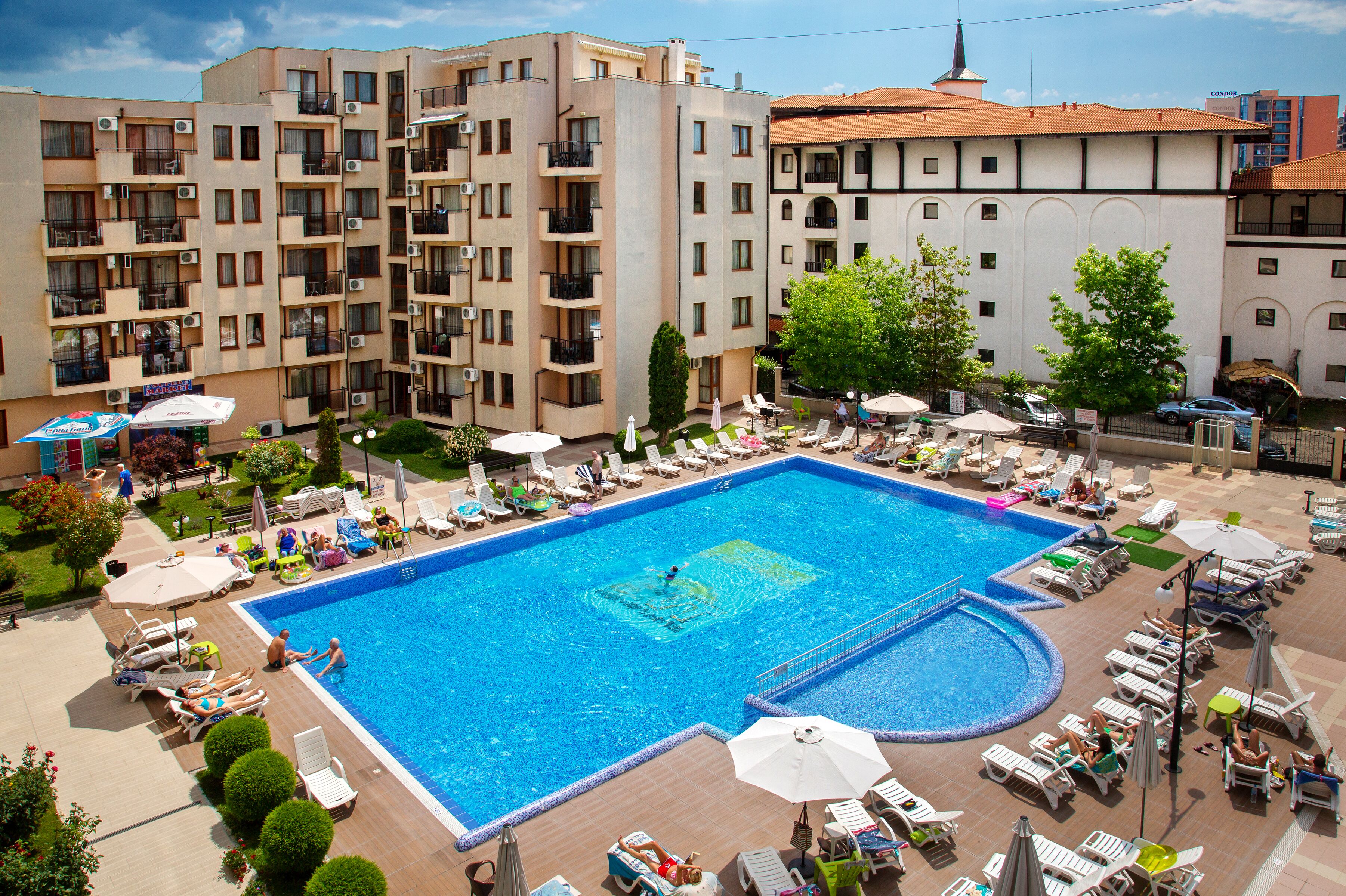 A large swimming pool surrounded by lounge chairs and umbrellas, with a multi-story building in the background.
