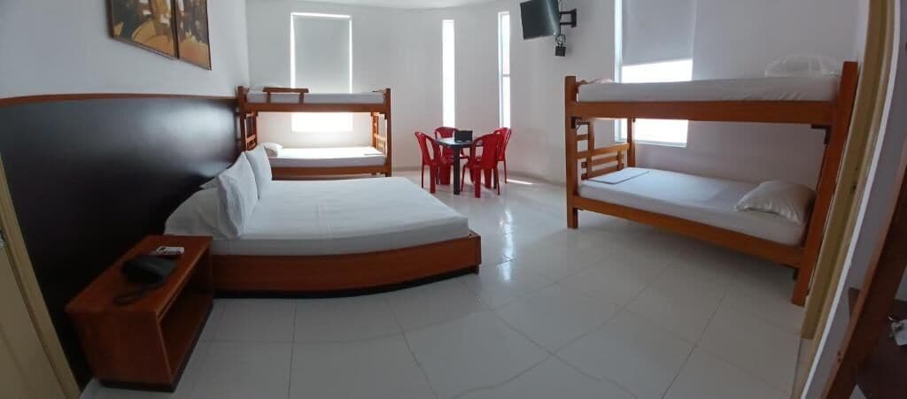 A room with a large bed, two bunk beds, a desk with chairs, and a TV on the wall.