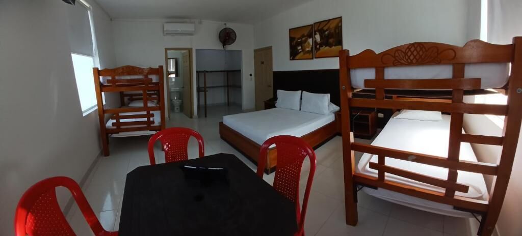 Una habitación con literas, una mesa y sillas. Hay una cama con cabecera de madera y una cama más pequeña con colchón blanco. En el centro, una mesa con mantel negro y sillas rojas.