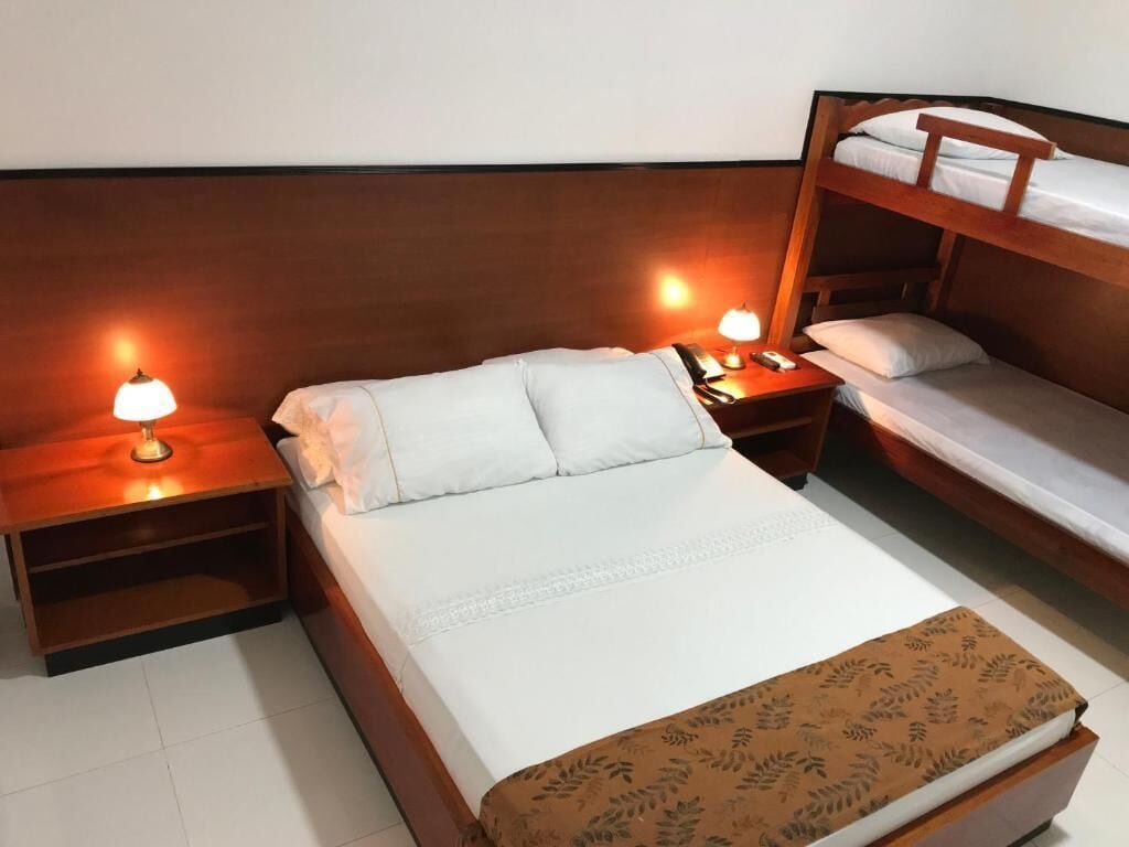 Una habitación de hotel con una cama individual, una cama litera, dos mesitas de noche con lámparas y una manta decorativa.