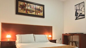 Une chambre d’hôtel bien aménagée, avec un grand lit, deux lampes de chevet, une tête de lit en bois, un bureau avec une chaise et deux œuvres d’art encadrées accrochées aux murs.