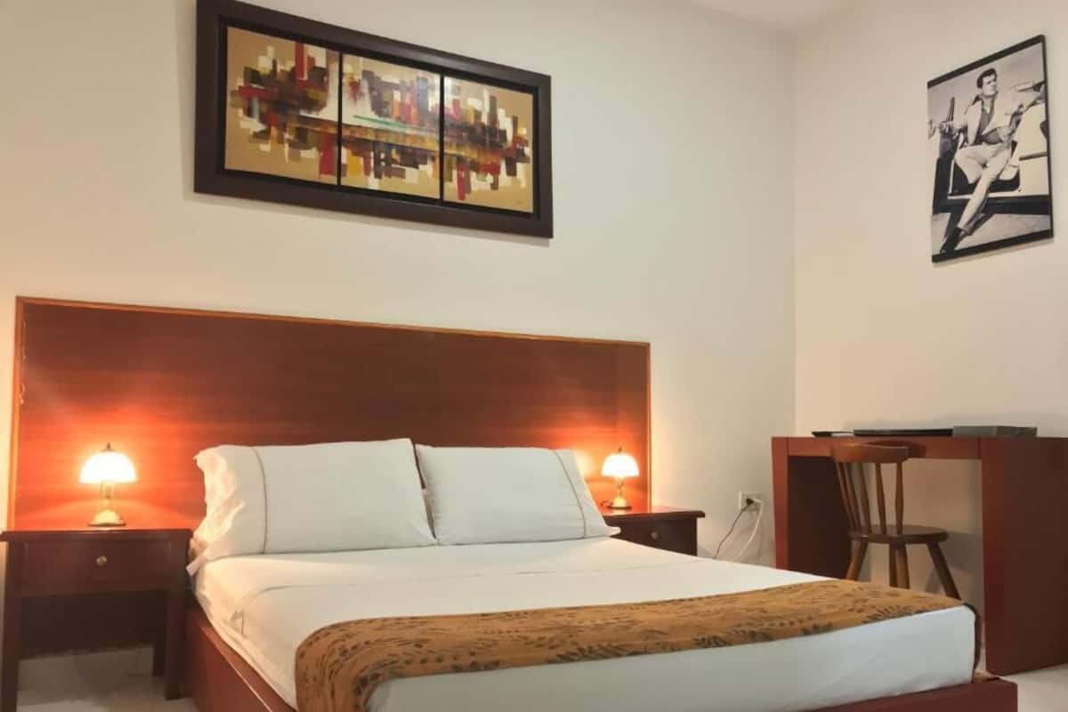 Habitación de hotel ordenada con una cama grande, dos lámparas de noche, cabecera de madera, escritorio con silla y dos obras de arte enmarcadas en las paredes.