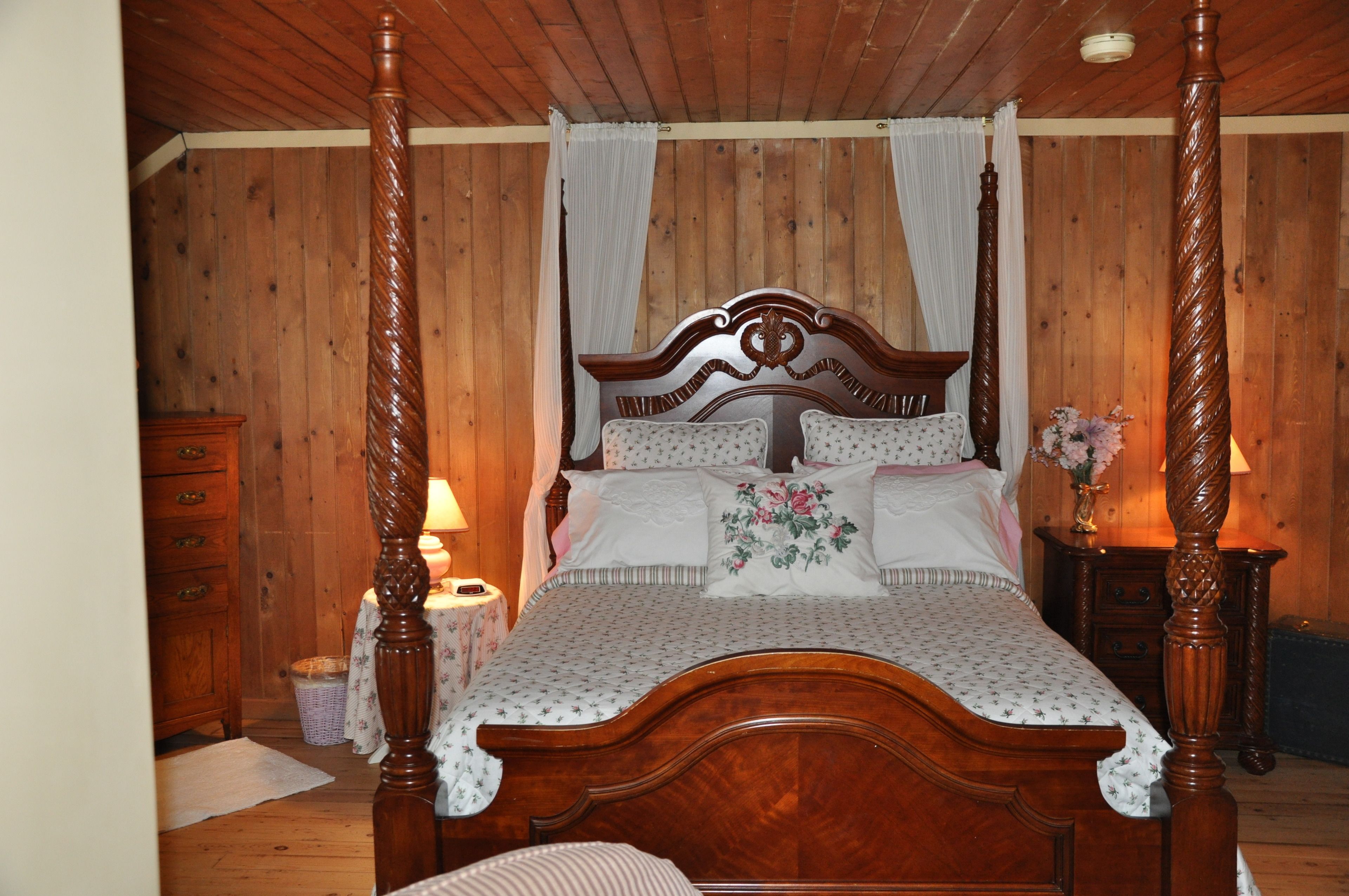 Een hemelbed met houten hoofdeinde, een nachtkastje met een lamp en een bijzettafeltje met een bloemenvaas.