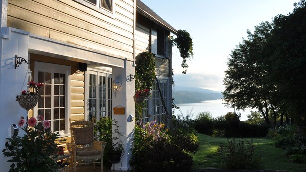 Ein zweistöckiges Haus mit Kiesweg, einer Veranda mit einem Stuhl und Blick auf einen See.