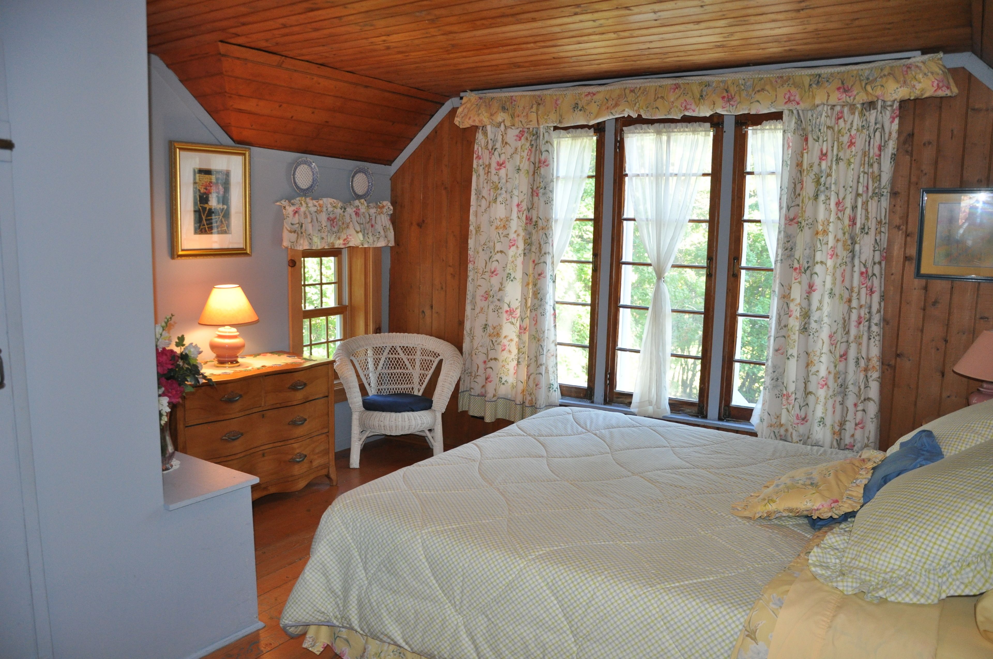 A cozy bedroom with a wooden ceiling, a bed with floral bedding, a chair, and a dresser with a lamp.